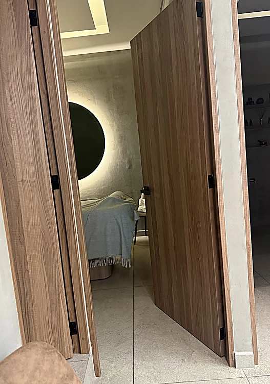Puerta de nogal abierta a la cabina de faciales y masaje, con camilla y un espejo redondo con luz de contorno