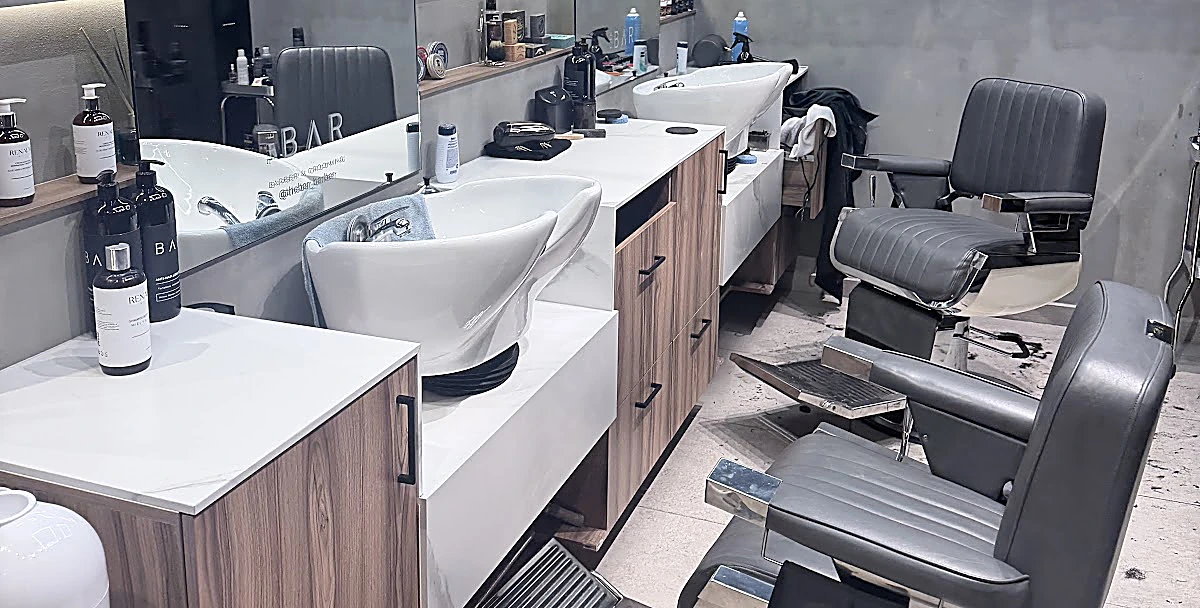 Estaciones de The Bar: lavabos blancos sobre muebles de nogal, sillones grises con reposapiés cromado y muro de concreto