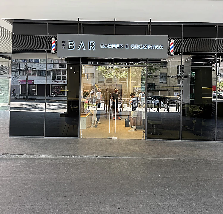 Fachada de vidrio de The Bar con el letrero iluminado y dos postes de barbero, bajo el plafón de lamas del edificio