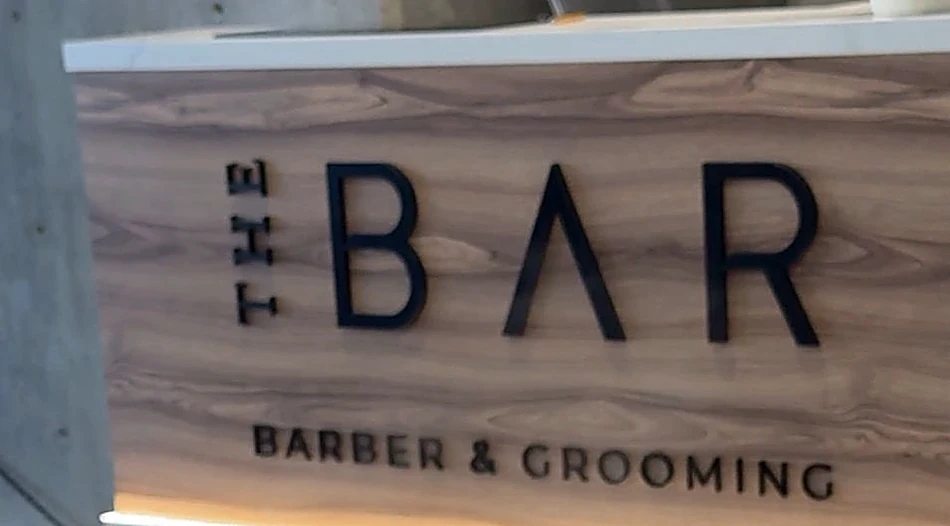 Panel de nogal de la recepción con el logo THE BAR, BARBER & GROOMING en letras negras y una tira de luz cálida